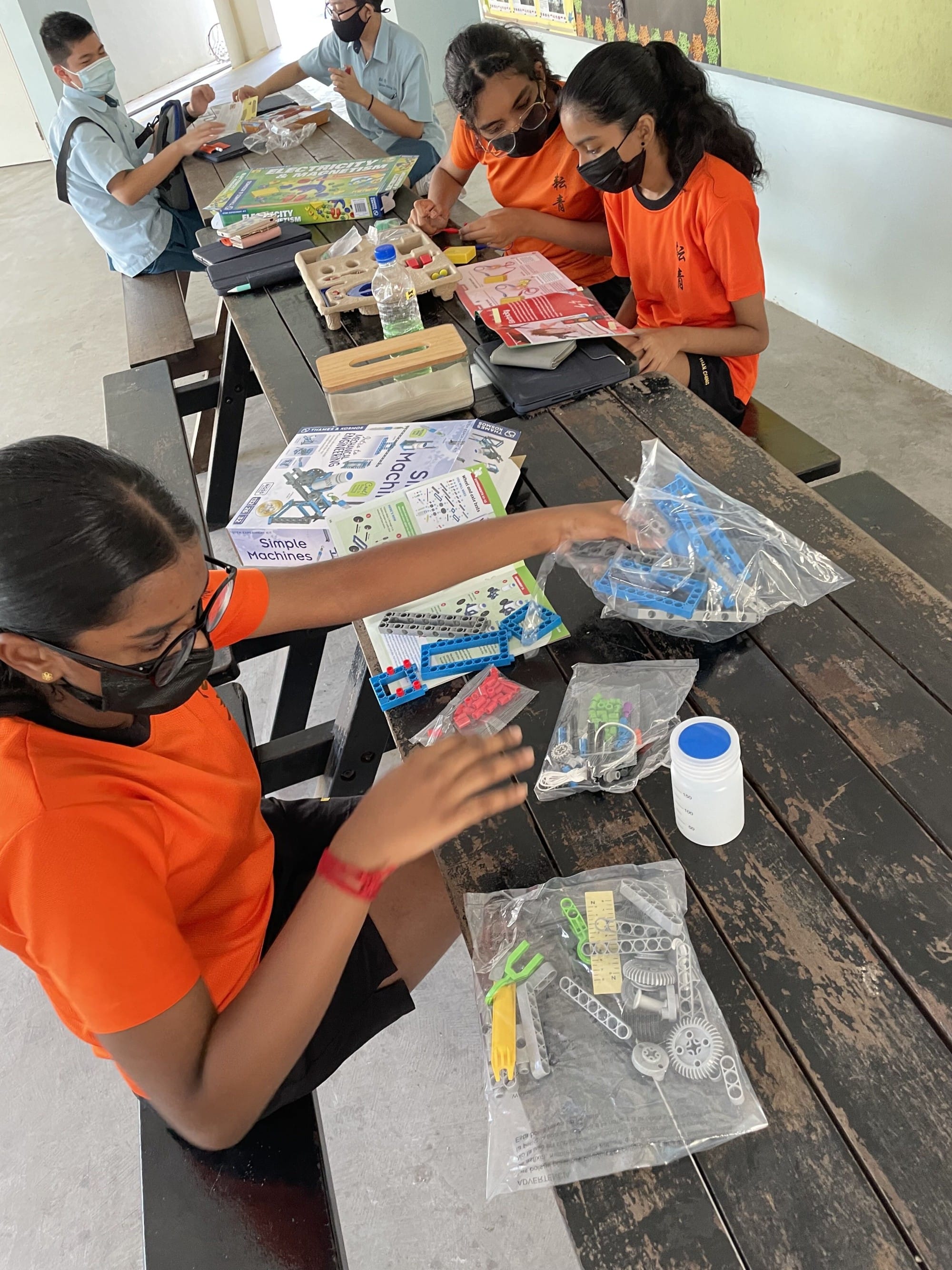 Students collaborate at an outdoor table assembling simple machine kits and learning from instructional materials during a hands-on science activity.